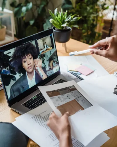 An HR interviewer reviewing printed notes at a desk while a candidate speaks on a laptop video call.