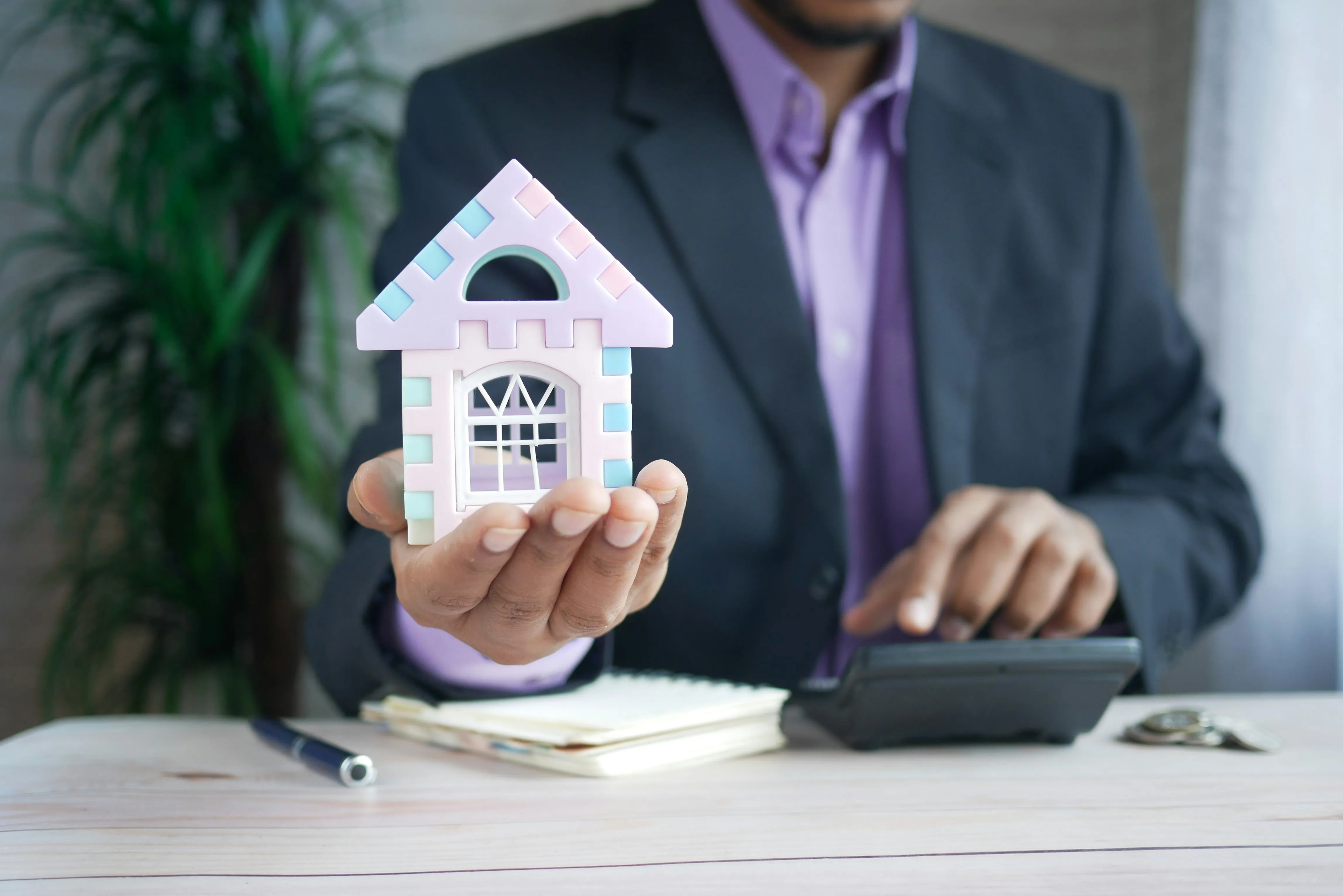 A man in a suit holding a small model house in one hand while working a calculator with the other.