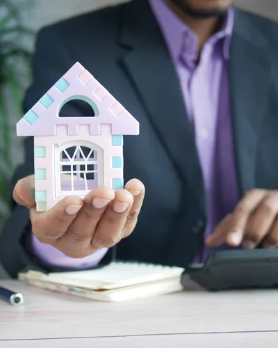 A man in a suit holding a small model house in one hand while working a calculator with the other.