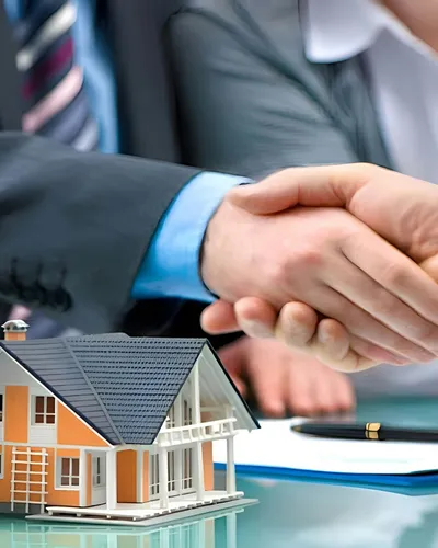 Two people in business attire shaking hands over a desk, with a model house and a set of keys nearby.