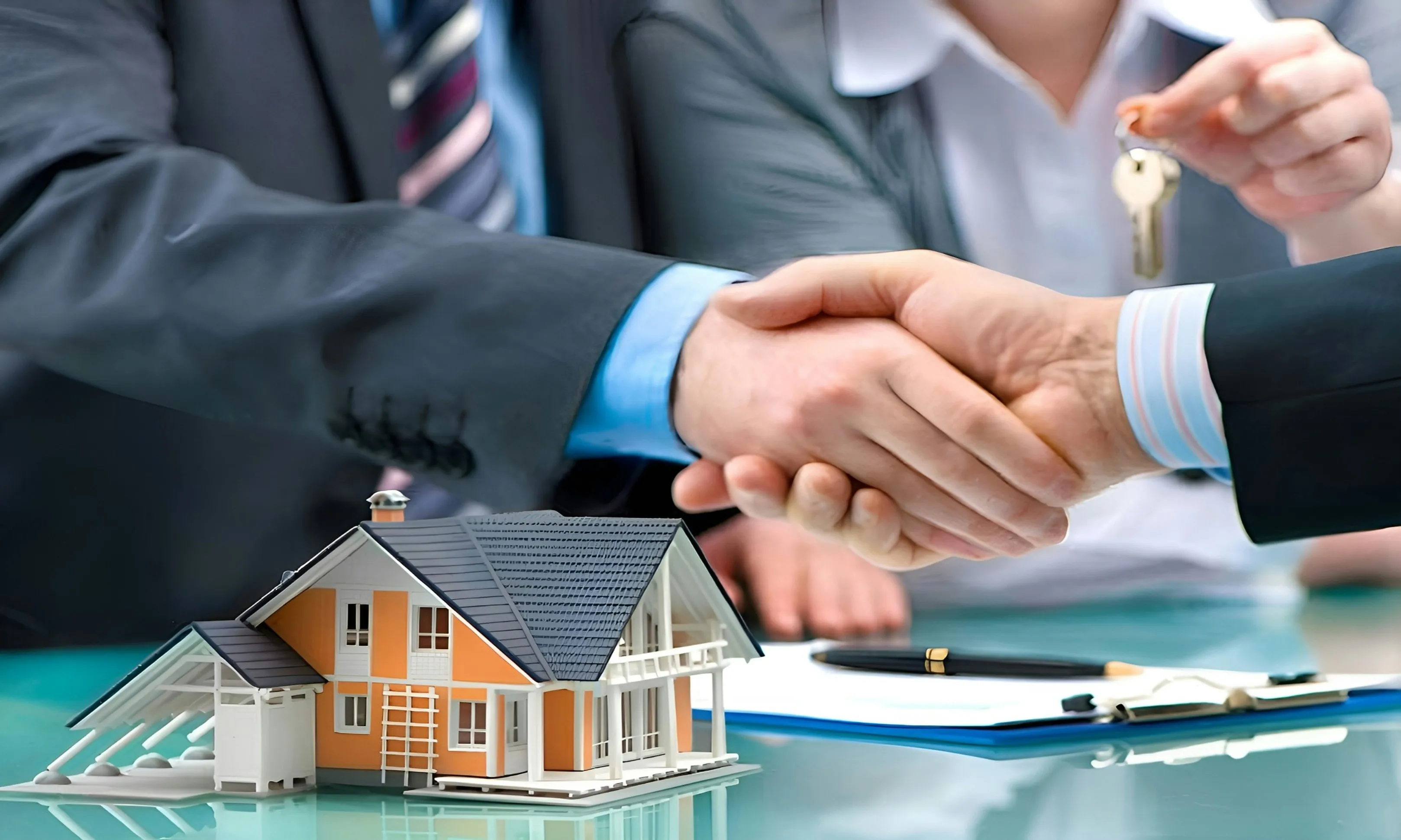 Two people in business attire shaking hands over a desk, with a model house and a set of keys nearby.